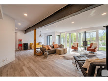 Load image into Gallery viewer, The image displays the exterior of the Wollf House, a large estate with a modern architectural design, set in a scenic location with trees and a body of water in the background.