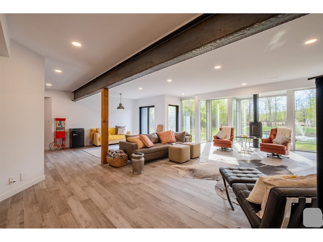 The image displays the exterior of the Wollf House, a large estate with a modern architectural design, set in a scenic location with trees and a body of water in the background.